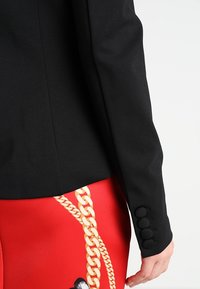 Veste noire sur mesure avec trois boutons ronds sur le poignet, associée à un pantalon rouge arborant un motif de chaîne dorée.