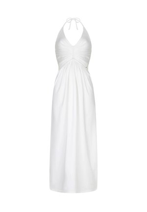 Woman in white halter maxi dress and sandals standing outdoors near cushioned seating and hanging wicker lamp in a sunny garden setting.