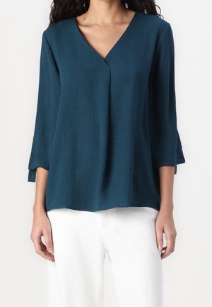Femme portant un chemisier bleu sarcelle à col en V avec des manches trois-quarts et un pantalon blanc, debout devant un fond clair uni.