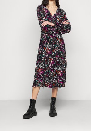 Femme portant une robe midi noire à imprimé floral coloré et manches longues, associée à des bottes de combat noires à lacets, debout les bras croisés.