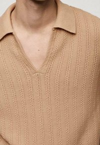 Vaaleanbeige neulepolo, jossa on V-pääntie, ja se on varustettu ribbikuvioinnilla ja pystysuorilla raidoilla. Kaulus on litteä ja pehmeä, mikä korostaa rentoa tyyliä.