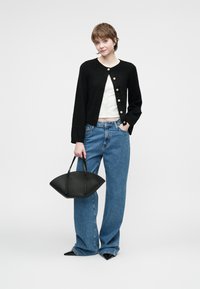 Černý pletený cardigan se zlatými knoflíky, spárovaný s bílým topem a modrými džíny s vysokým pasem. Modelka drží černou, strukturovanou kabelku.
