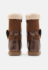 Bottines en suède marron avec doublure en fausse fourrure, accents en cuir et couture décorative. Comprend une sangle marron et des fermetures métalliques.