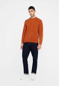 Oranger gestrickter Pullover mit strukturiertem Muster, rundem Halsausschnitt und langen Ärmeln, kombiniert mit dunkelblauen Jeans und weißen Sneakers.