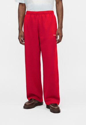 Isik, kes kannab säravpunaseid laia säärega pükse elastse vöökohaga, pruune loafers-kingi ja valget särki, seisab ühevärvilise tausta ees.