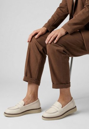 Person iført brune opsmøgede bukser og cremefarvede loafers, siddende med hænderne hvilende på knæene mod en ensfarvet grå baggrund.