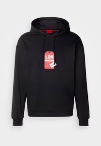 Sweat à capuche noir avec une étiquette rouge à l'avant indiquant « LDN », « Fragile » et une petite étiquette blanche portant l'inscription « Hug ».