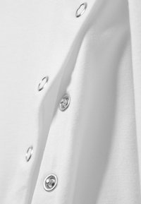 Weißes Baumwollhemd mit silbernen Druckknöpfen entlang der Naht, mit glatter Textur und einem eleganten, minimalistischem Design.