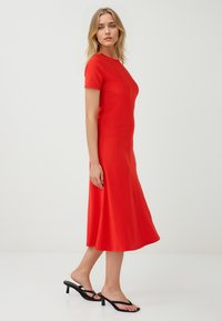 Een vrouw staat zijwaarts gekeerd in een strak rood jurkje en zwarte sandalen, wat een stijlvolle uitstraling geeft tegen een effen witte achtergrond.