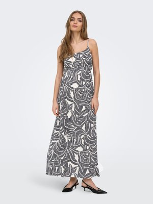 Femeie purtând o rochie maxi neagră cu alb, fără mâneci, cu model cu frunze, bretele subțiri și tocuri slingback negre, stând în fața unui fundal simplu.