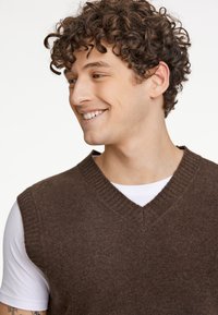 Brun stickad väst med V-ringning och ribbad krage, bärs över en vit t-shirt. Har en mjuk textur och inga synliga mönster eller detaljer.