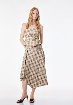 Femme portant un top bustier à carreaux beige et marron avec un nœud et une jupe portefeuille asymétrique assortie, portée avec des sandales à talons marron foncé.