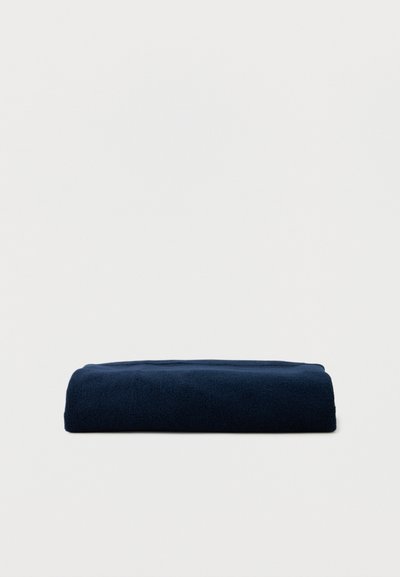 Donkerblauwe opgevouwen handdoek geplaatst op een effen lichtgekleurde ondergrond, met zichtbaar zachte stoftextuur en scherpe randen.