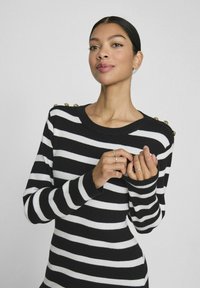 Pull rayé noir et blanc avec un col rond, des manches longues et des accents de boutons dorés sur l'épaule. Texture côtelée sur l'ensemble.