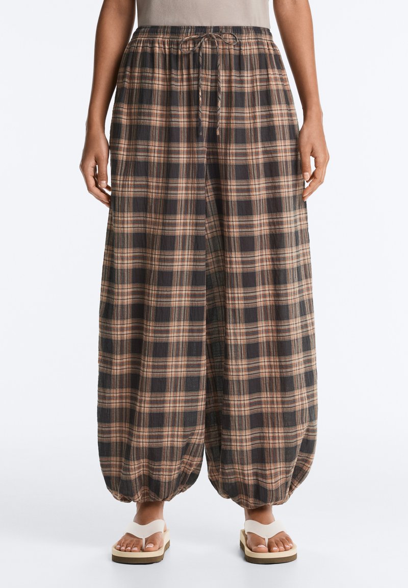 Pantalon ample à carreaux avec poignets élastiques aux chevilles et taille à cordon, porté avec des sandales compensées blanches.