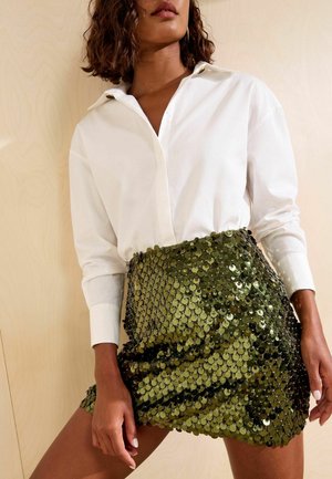 Mujer con una camisa blanca de manga larga metida en una minifalda verde oliva con lentejuelas sobre un fondo de madera clara.