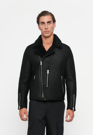 Jeune homme portant une veste en shearling noire avec des fermetures éclairs argentées et un pantalon cargo noir, se tenant contre un fond blanc.