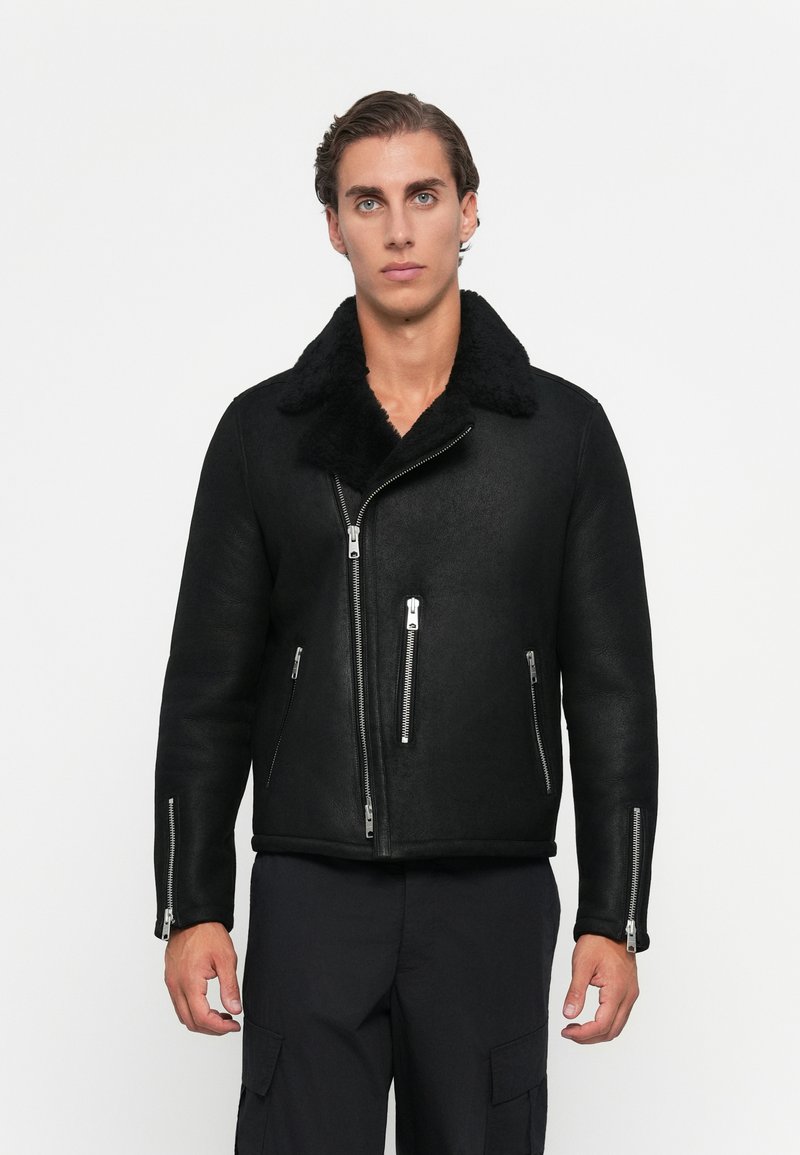 Young man wearing a black shearling jacket with silver zippers and black cargo pants standing against a white background.