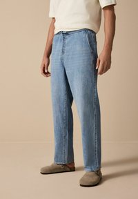 Jeans din denim de culoare albastru deschis, cu croi drept și cusături contrastante; purtate cu pantofi slip-on bej, stilizate cu o cămașă albă.