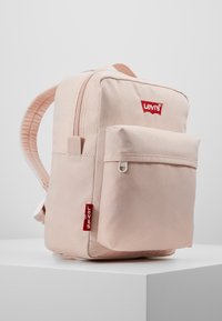 Vaaleanpunainen kangaskassi, jossa on etupaneelissa vetoketjullinen tasku, pehmustetut olkaimet ja punainen Levi's-logo sekä edessä että sivulla.