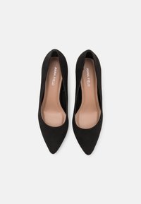 Chaussures à talons hauts en suede noir avec un bout pointu et un design ouvert sur le côté. L'intérieur présente une semelle intérieure beige clair avec le logo "ANNA FIELD".