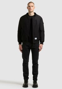 Czarna kurtka bomberka z kieszenią na zamek i zatrzaskami, noszona na czarnym swetrze, zestawiona z ciemnymi dżinsami i czarnymi butami.