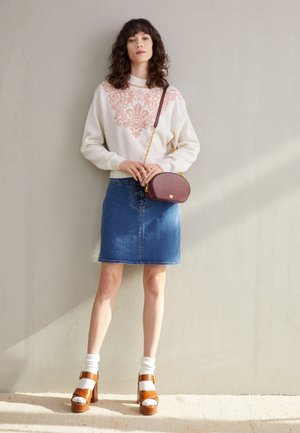 Femme portant un pull blanc avec un motif rose, une jupe en jean bleue, des sandales marron à talons avec des chaussettes blanches, et portant un petit sac marron.