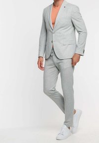 Mężczyzna w jasnoszarym żakiecie i dopasowanych spodniach dresowych, w pomarańczowej koszuli i białych trampkach, stoi na białym tle.