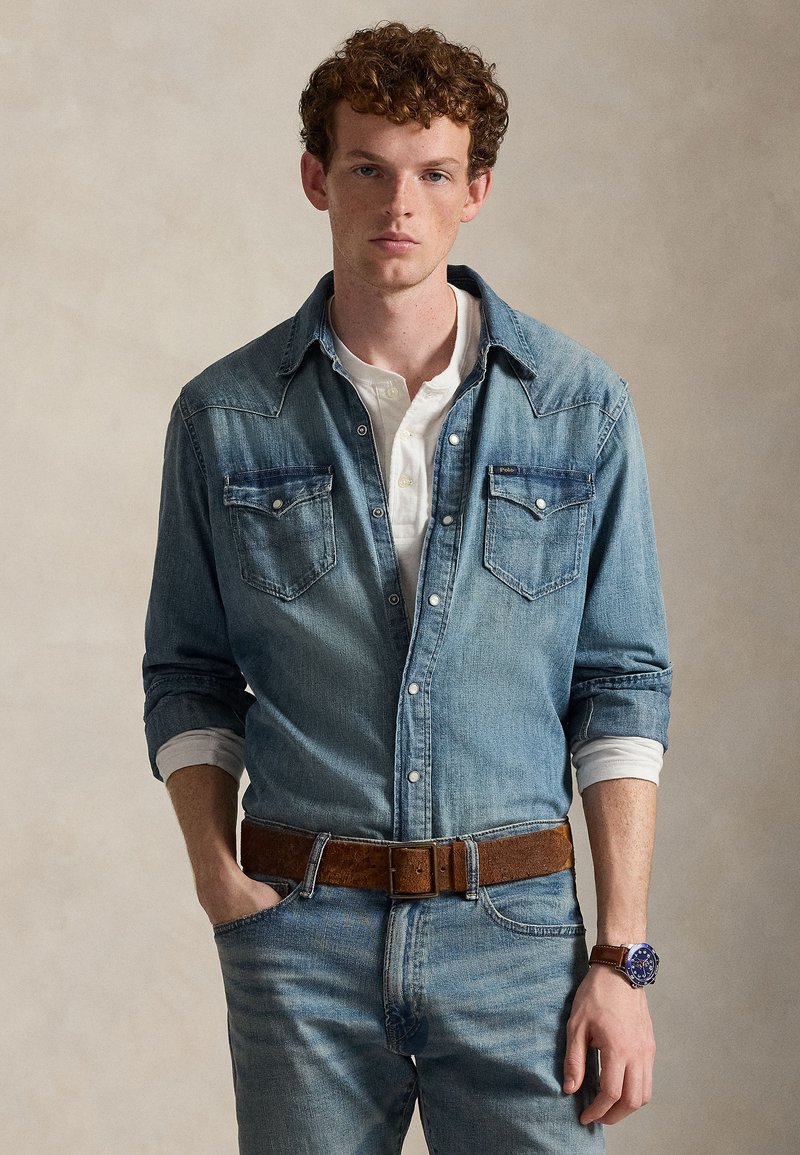 Jonge man met krullend haar, die een denimoverhemd draagt over een witte henley, blauwe spijkerbroek, een bruine riem en een bruine leren horloge.