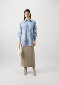 Camicia a righe blu e bianche con taschino frontale, abbinata a una gonna di lunghezza al ginocchio color marrone chiaro e stivaletti beige, mentre tiene una borsa bianca.