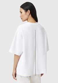 Witte oversized short-sleeve shirt met een centrale rits aan de achterkant en een gladde textuur, met een relaxte pasvorm en minimalistisch ontwerp.
