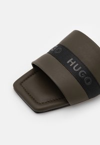 Sandale à talon ouvert de couleur vert olive avec une sangle noire présentant le branding en relief "HUGO" sur le dessus.
