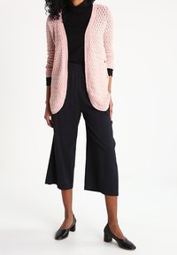 Cárdigan de punto abierto en rosa claro sobre un cuello de tortuga negro, combinado con pantalones cortos negros y zapatos de tacón negros. Texturas suaves y corte holgado.
