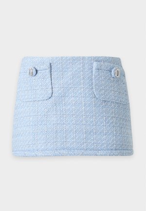 Femme debout portant une veste texturée bleu clair et une jupe assortie avec des poches avant, un T-shirt graphique blanc et des chaussures noires pointues.
