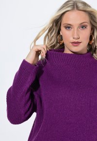 Lila gestrickter Pullover mit einem hohen, umgeschlagenen Kragen und gerippter Textur. Das Modell hält den Kragen mit einer Hand und zeigt die Weichheit des Stoffes.