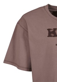 Brown cotton t-shirt with a crew neckline. Features short sleeves and a slightly relaxed fit. Front displays a faded black logo.