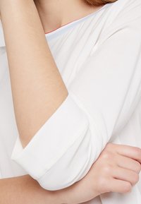 Camicia bianca con scollatura rotonda e dettagli blu e rossi. Le maniche sono arrotolate, mettendo in evidenza un tessuto leggero e morbido.