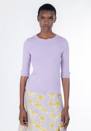 Kvinde iført en lavendelfarvet top med opkrogede ærmer og guldknapper, kombineret med en lavendelfarvet nederdel med gule blomsterprint.