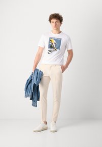 Menns antrekk med en hvit t-skjorte med sykkeldiagram, kremfargede slim-fit bukser, blå denimjakke og hvite joggesko.