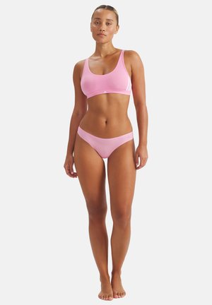 Frau steht barfuß, trägt einen hellrosa Sport-BH und passende Bikini-Unterwäsche vor einem schlichten weißen Hintergrund.