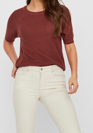 Femme portant un top tricoté à manches courtes couleur rouille, rentré dans un pantalon ajusté taille haute beige clair, sur un fond uni.