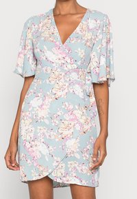 Květinové zavinovací šaty v světle modré barvě s krátkými volánovými rukávy, růžovými a bílými květinovými vzory a pevně utaženým páskem.