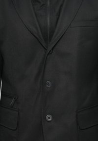 Svart blazer med en slät tygtextur, slagkrage, en-knapps stängning och två framfickor med dragkedjedetalj.
