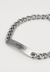 Sølv metalkjedearmbånd med en flat plate gravert med "Ed Hardy." Lenkene er ovale og polerte for en blank finish.