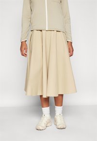 Beige midi szoknya, teltebb sziluettel, könnyű anyagból készült. Zippzáros felsővel és világos színű sportcipővel, fehér zoknival párosítva.