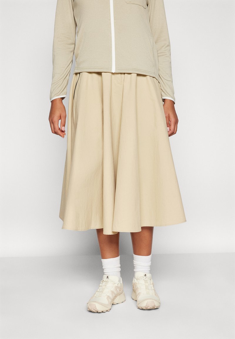 Beige midi szoknya, teltebb sziluettel, könnyű anyagból készült. Zippzáros felsővel és világos színű sportcipővel, fehér zoknival párosítva.