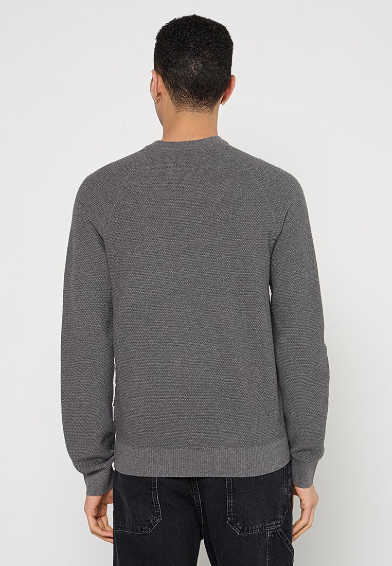 Person von hinten gesehen, der einen strukturierten grauen langärmligen Pullover und schwarze Jeans trägt und vor einem schlichten weißen Hintergrund steht.