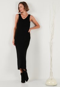 Black ribbed knit sleeveless dress with a V-neck, fitted shape, and side slits. Paired with black ankle boots featuring gold hardware accents.