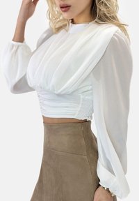 Hvid bluse med puff-ærmer og rynket overdel, kombineret med en beige ruskindnederdel med sidelukning. Glat tekstur og elegant design.