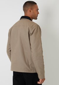 Beige katoenen jack met een zwarte corduroy kraag, eenvoudig ontwerp en lange mouwen. Geen zichtbare patronen of hardwaredetails.